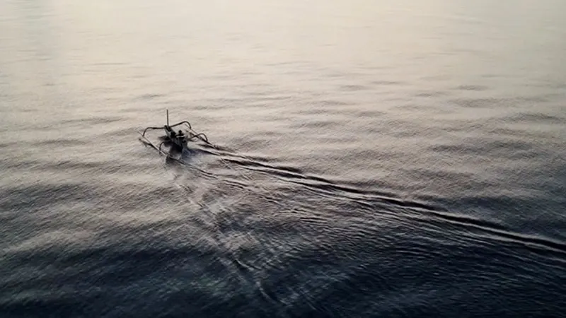A jukung fishing boat seen from above, its wake trailing across calm open water off the Tianyar coast