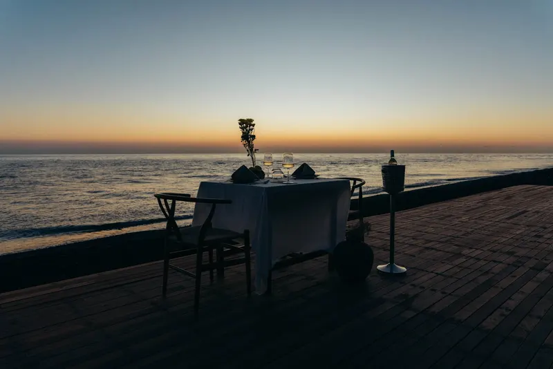 A table laid with white cloth and wine glasses on the deck at the water's edge, the sun already down and the horizon still warm
