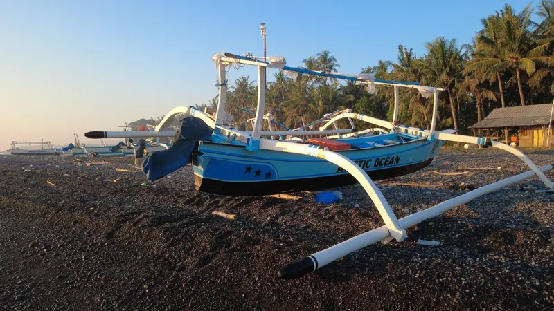 A jukung outrigger fishing boat resting on the pebble shore at Tianyar in early morning light, with palm trees and a small shore hut behind.