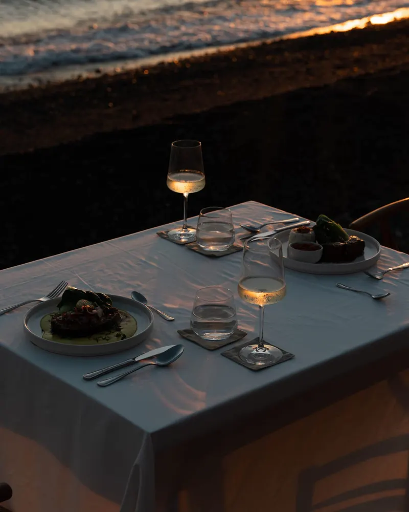 A laid table with a plate and a glass of wine on the deck at the shore, the pebbles and the sea in the low sun