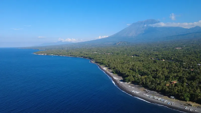Aerial view of Bali's north-east coastline curving towards Mount Agung, seen across the sea on a clear day with light cloud around the summit.