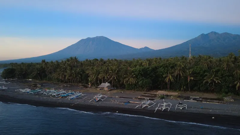 Aerial view of the Tianyar shore at dusk, fishing boats and a palm treeline along the coast with the mountains behind.