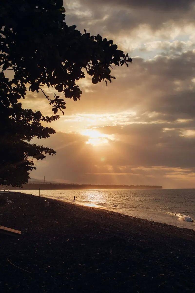 The sun breaking through cloud over the sea near Tianyar, a figure walking along the shore in the evening light