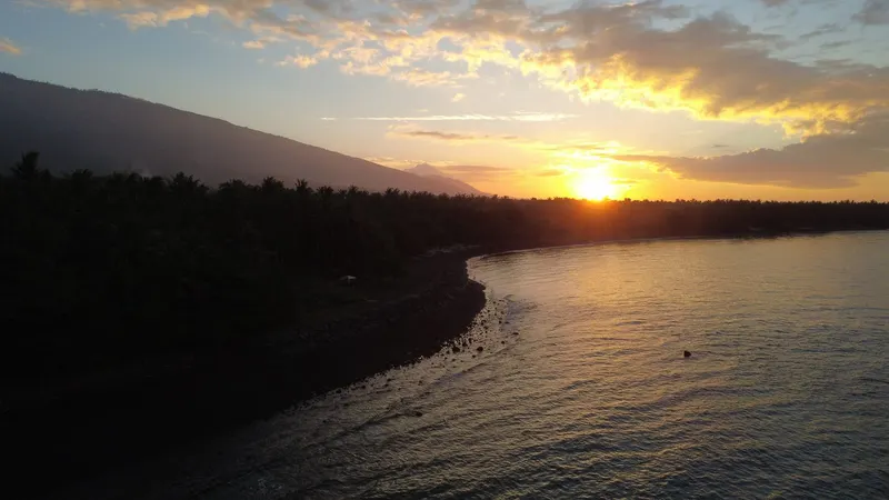 The sun setting into cloud over the sea at Tianyar, silhouetted palm trees and a dark pebble shore in the foreground.