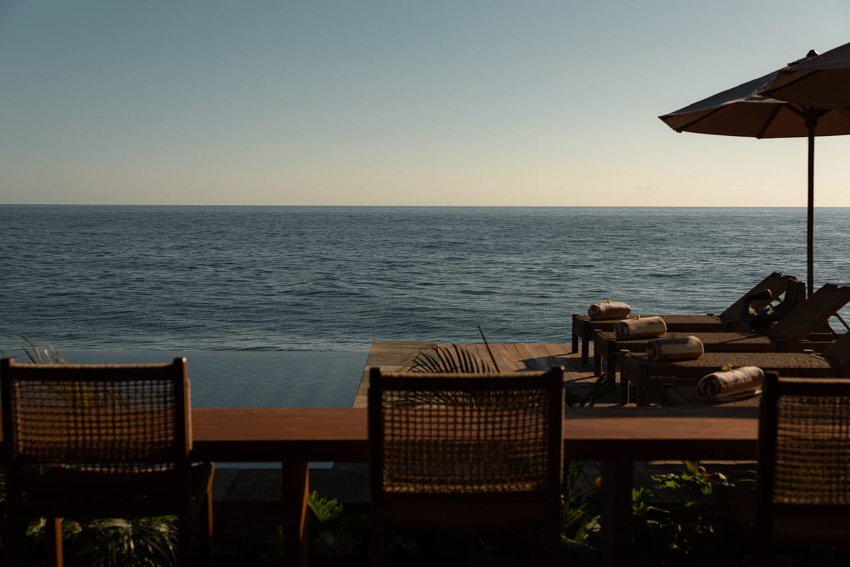 Wicker chairs and a table on the timber deck by the sea at Tianyar, the horizon in soft evening light